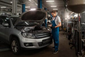 A mechanic in blue coveralls inspects the engine of a silver SUV with the hood open inside an auto repair shop.